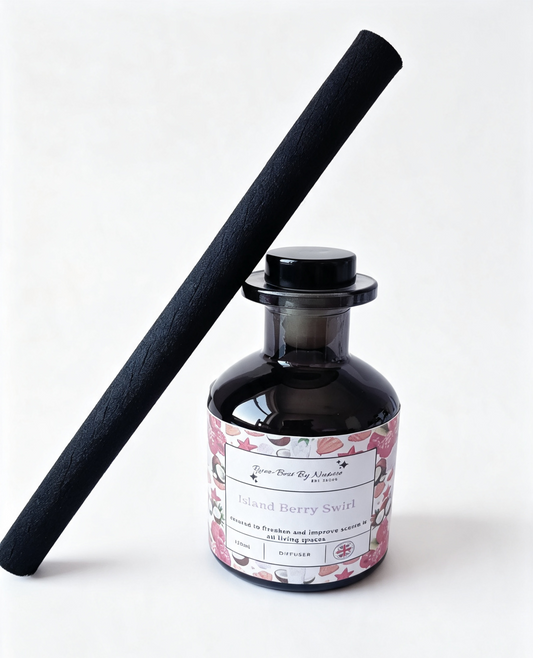 A reed diffuser bottle with a black glass body and a floral pink label, alongside a tall black reed stick, on a clean white background.