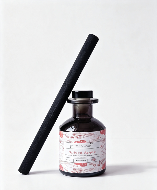 A dark amber glass reed diffuser bottle with a black cap and a long black reed leaning diagonally against it. The bottle has a white label with red and pink botanical illustrations and the text 'Spiced Apple'