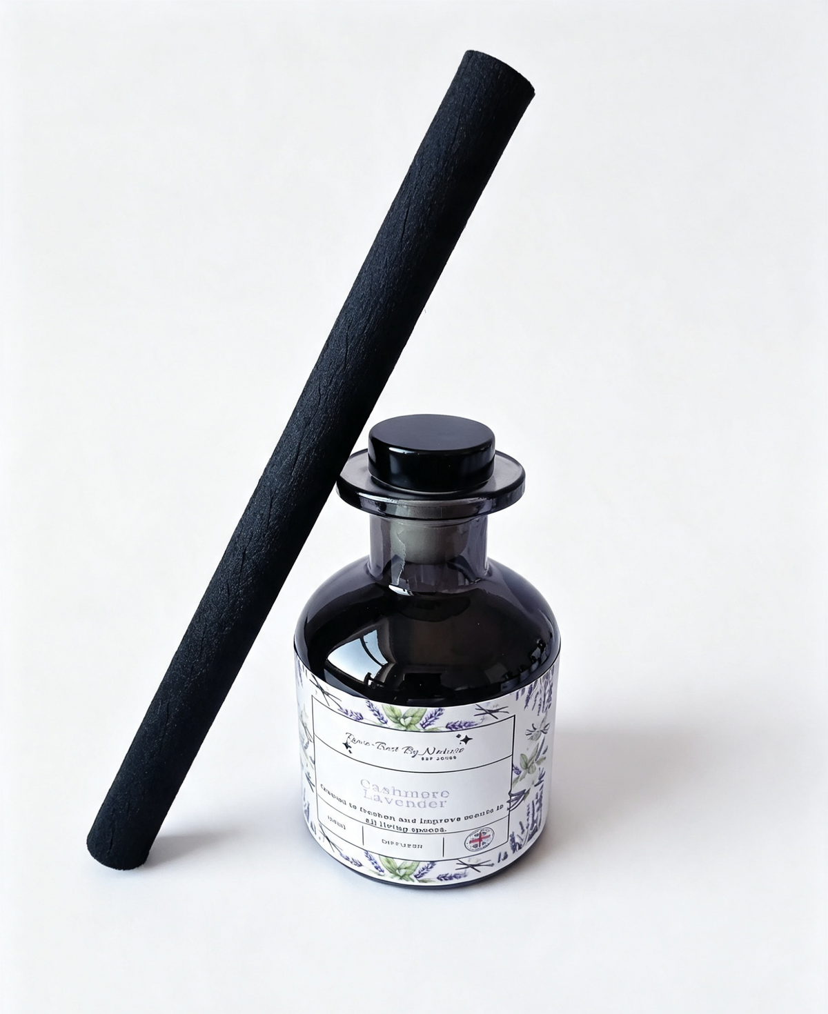 A reed diffuser bottle with a black glass body and a white botanical label, standing next to a long black reed stick on a clean white background.