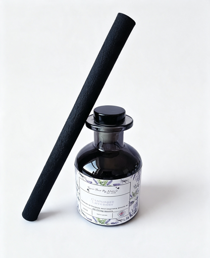 A reed diffuser bottle with a black glass body and a white botanical label, standing next to a long black reed stick on a clean white background.