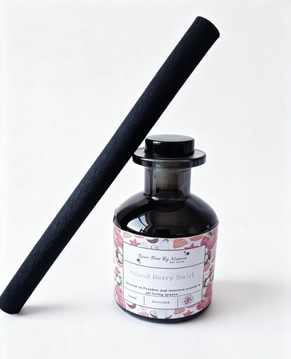 A reed diffuser bottle with a black glass body and a floral pink label, alongside a tall black reed stick, on a clean white background.