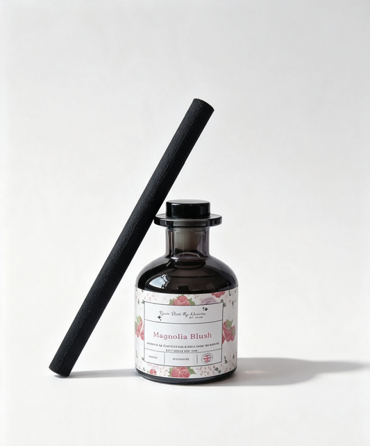 A dark amber glass reed diffuser bottle with a floral label reading 'Magnolia Blush' and a black reed stick leaning diagonally against it on a white background.