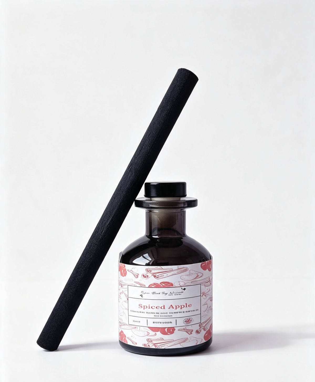 A dark amber glass reed diffuser bottle with a black cap and a long black reed leaning diagonally against it. The bottle has a white label with red and pink botanical illustrations and the text 'Spiced Apple' 