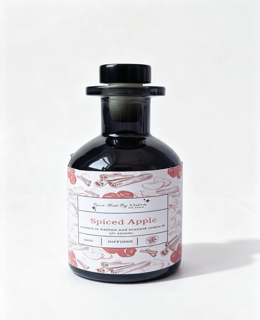 A dark glass diffuser bottle with a black cap and a white label featuring pink illustrations of cinnamon sticks and red apples, labeled 'Spiced Apple' by Rowe-Best By Nature
